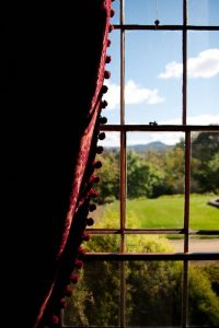 A pane glass window with a red brocade curtain that has bobbles on the edging, overlooking a green verdant lawn and garden