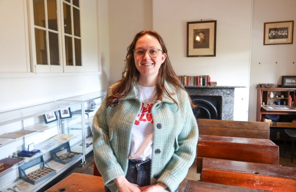 A smiling young woman with brown hair and glasses stands in front of old school desks.