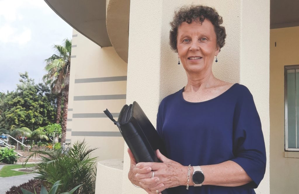 A woman with dark grey curly hair in a navy top holding a folder.