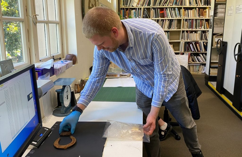 A smiling young man wearing a blue striped shirt and gloves examines an archaeological object.