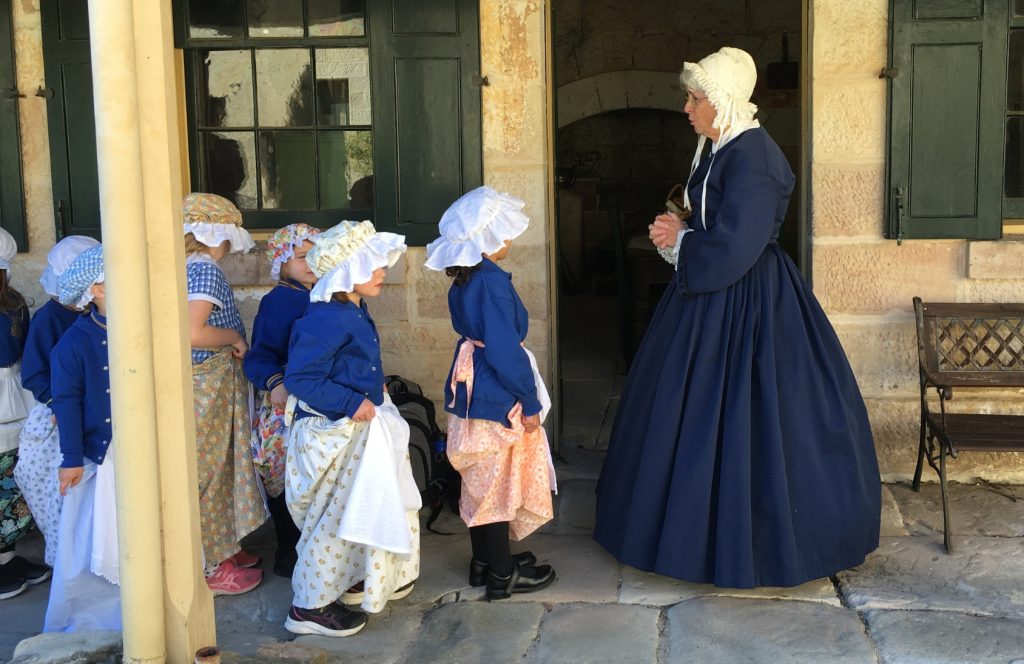 A lady in a bonnet and old-fashioned dress talks to school children.