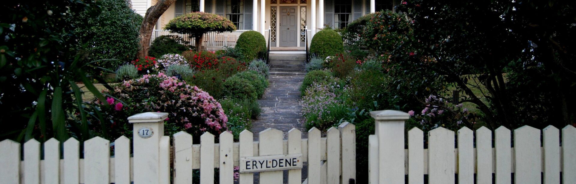 A white picket fence in front of a flowering garden and heritage house