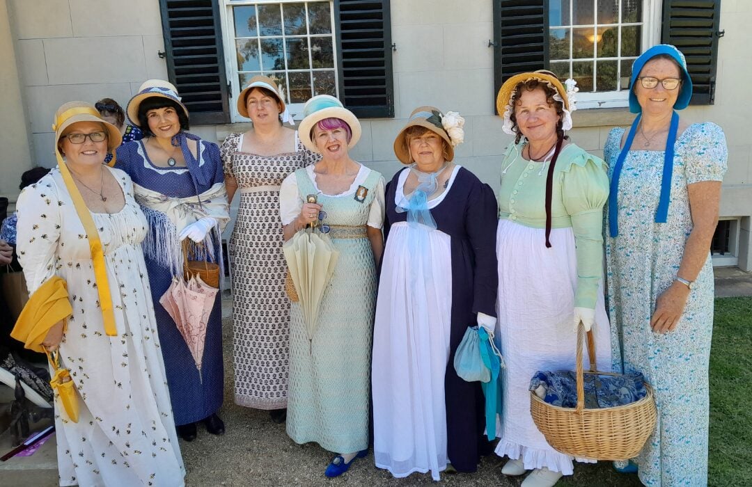 Women dressed in regency attire in front of a heritage building.