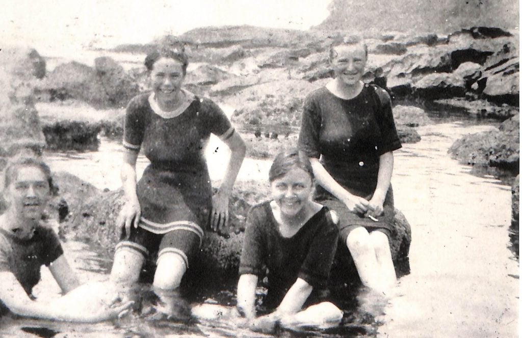 A black and white photograph of a group of girls in their late teens in vintage bathing suits pictured in rock pools at a beach.