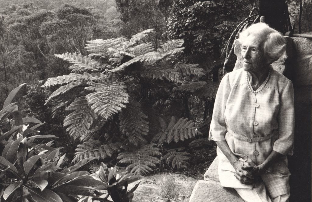 An elegantly dressed elderly woman pictured on a rock shelf against a background of ferns.
