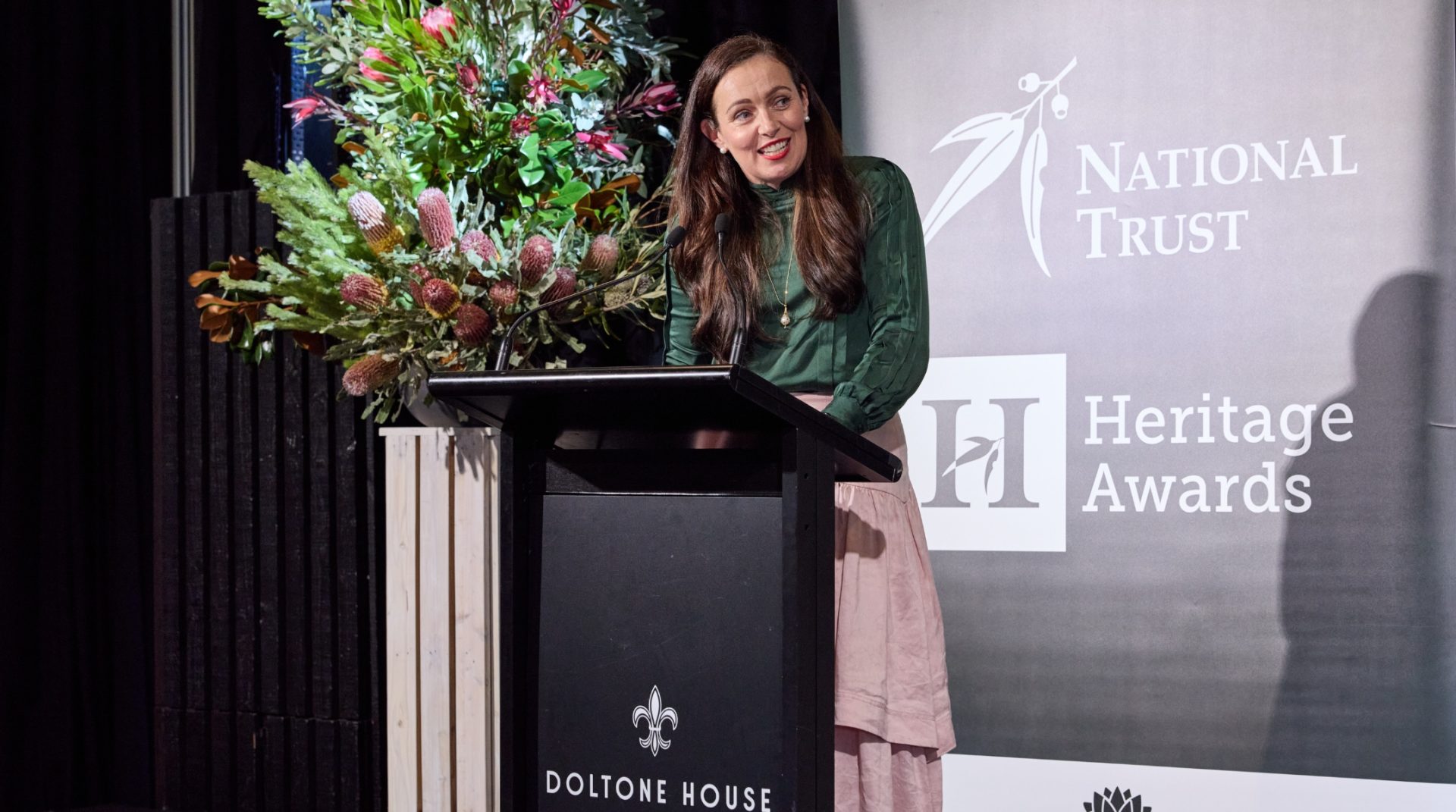 A lady with long dark brown hair, a green top and blush-pink skirt presents from a lectern.